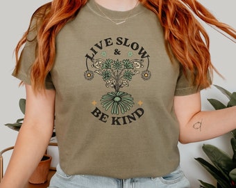May include: A khaki green t-shirt with a floral design and the text "Live Slow & Be Kind".