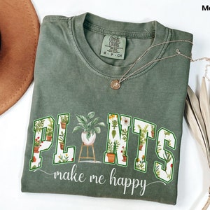 May include: A moss green t-shirt with the words "PLANTS make me happy" in a white and green design. The word "PLANTS" is filled with images of potted plants. A gold necklace and a brown hat are also in the image.