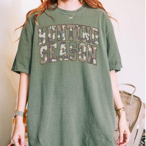 K&ouml;nnte beinhalten: Ein &uuml;berdimensioniertes T-Shirt in Moosgr&uuml;n mit einem Tarnmuster-Druck, auf dem "Hunting Season" steht.