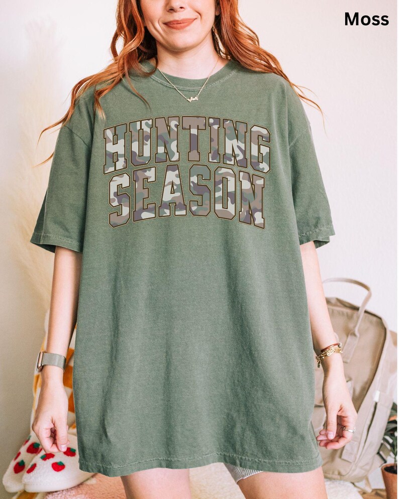 K&ouml;nnte beinhalten: Ein &uuml;berdimensioniertes T-Shirt in Moosgr&uuml;n mit einem Tarnmuster-Druck, auf dem "Hunting Season" steht.