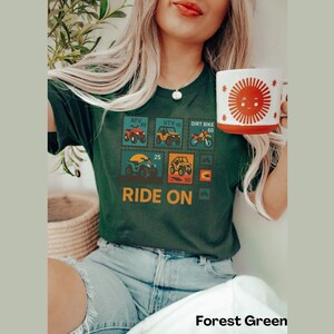 May include: Forest green t-shirt featuring a graphic of ATV, UTV, and dirt bike postage stamps with the words "RIDE ON". A white mug with a smiling sun design is held in the hand. The shirt is paired with blue jeans.