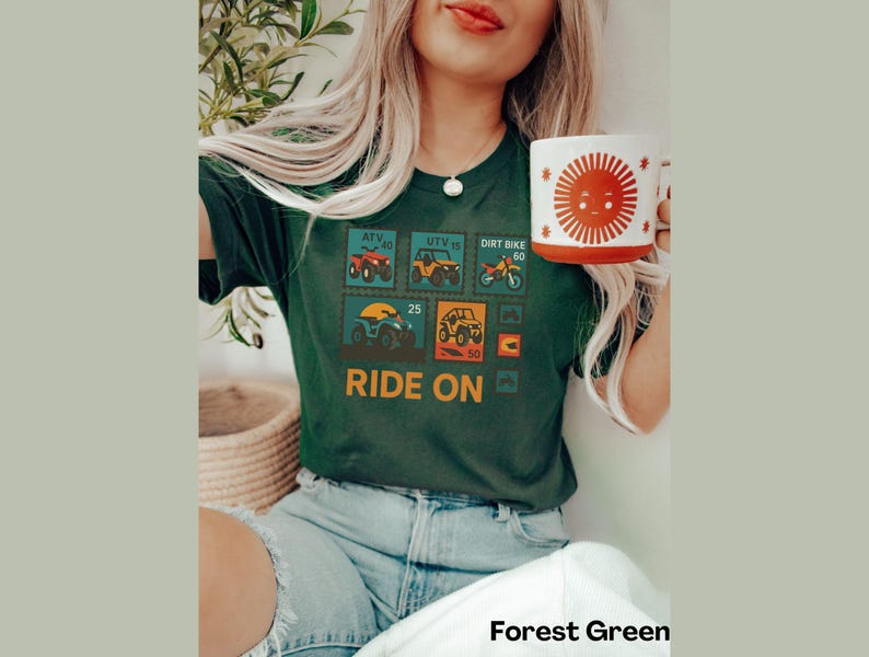 May include: Forest green t-shirt featuring a graphic of ATV, UTV, and dirt bike postage stamps with the words "RIDE ON". A white mug with a smiling sun design is held in the hand. The shirt is paired with blue jeans.