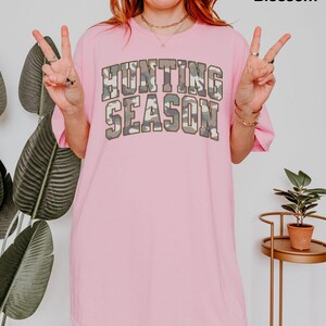K&ouml;nnte beinhalten: Rosafarbenes Oversize-T-Shirt mit dem Text "Hunting Season" in Camouflage-Druck.