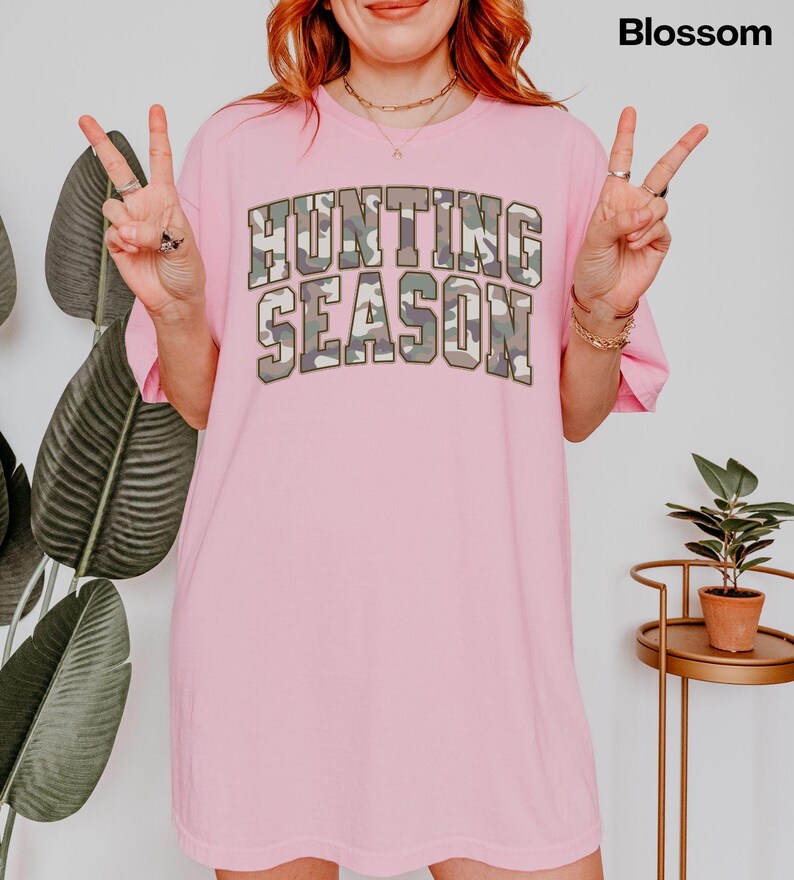 K&ouml;nnte beinhalten: Rosafarbenes Oversize-T-Shirt mit dem Text "Hunting Season" in Camouflage-Druck.