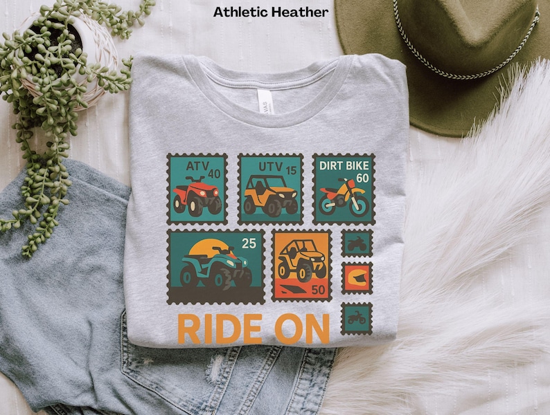 May include: Athletic Heather gray t-shirt with a graphic of ATV, UTV, and dirt bike postage stamps. The text "RIDE ON" is printed in orange. The shirt is made of a soft, comfortable material.