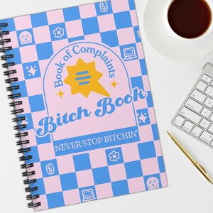 May include: A pink and blue chequered notebook with the text "Book of Complaints Bitch Book NEVER STOP BITCHIN'" in a retro font. The notebook has a spiral binding and a yellow speech bubble with three lines.