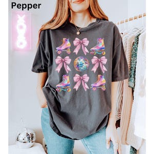 May include: A dark grey t-shirt featuring a graphic print of roller skates, pink bows, and a disco ball. The graphic is in a watercolour style with iridescent colours.