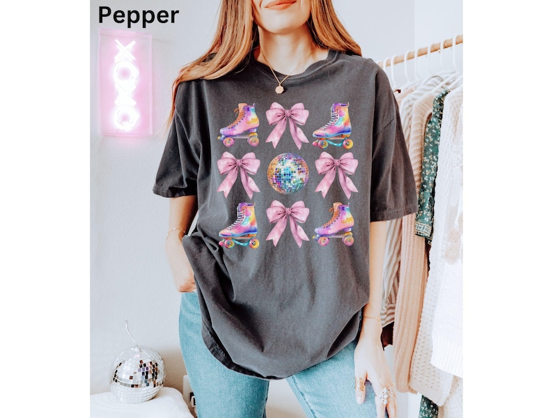 May include: A dark grey t-shirt featuring a graphic print of roller skates, pink bows, and a disco ball. The graphic is in a watercolour style with iridescent colours.