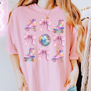 May include: A pink t-shirt with a graphic design featuring roller skates, disco balls, and bows. The roller skates are rainbow coloured and the bows are pink.