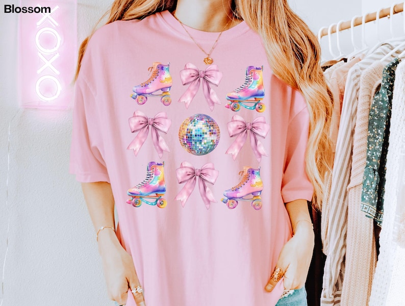 May include: A pink t-shirt with a graphic design featuring roller skates, disco balls, and bows. The roller skates are rainbow coloured and the bows are pink.
