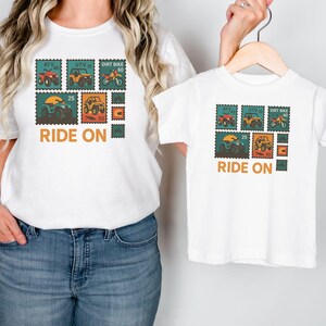 May include: White t-shirts with a stamp-like design featuring ATVs, UTVs, and dirt bikes in red, yellow, and orange. The text "RIDE ON" is printed below the stamps. One shirt is adult size, the other is child size.