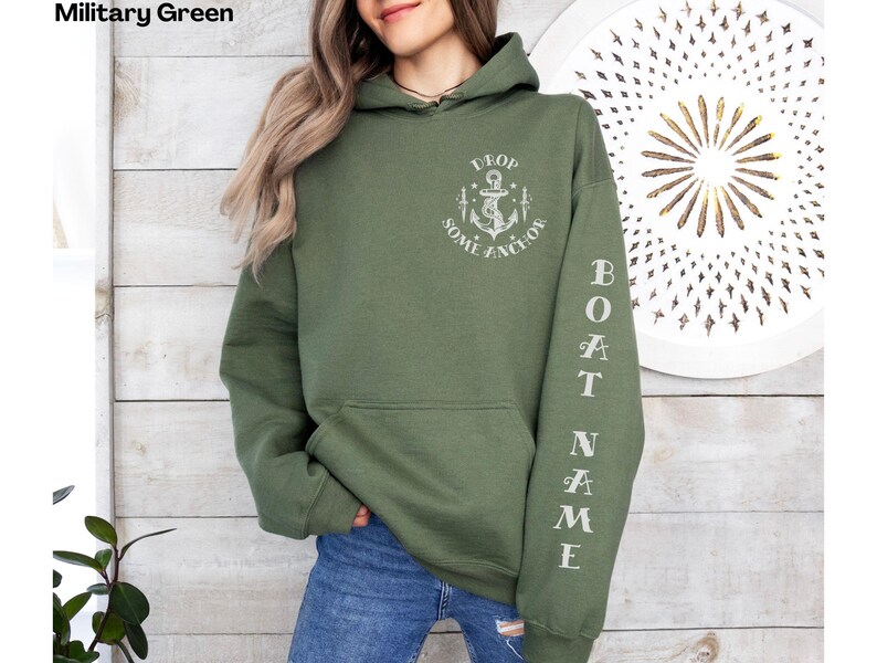 May include: A military green hooded sweatshirt with the text "DROP SOME ANCHOR" and an anchor graphic on the front. The left sleeve has the text "BOAT NAME" printed vertically.