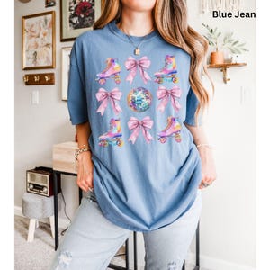 May include: A blue jean t-shirt featuring a graphic print of pink bows and roller skates with disco balls. The text "Blue Jean" is visible in the top right corner of the image.