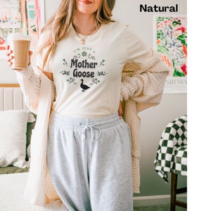 May include: Cream-colored t-shirt with the text "I'm Just a Mother Goose" in a floral design, featuring a black goose silhouette. The shirt is paired with gray sweatpants and a cream-colored cardigan.