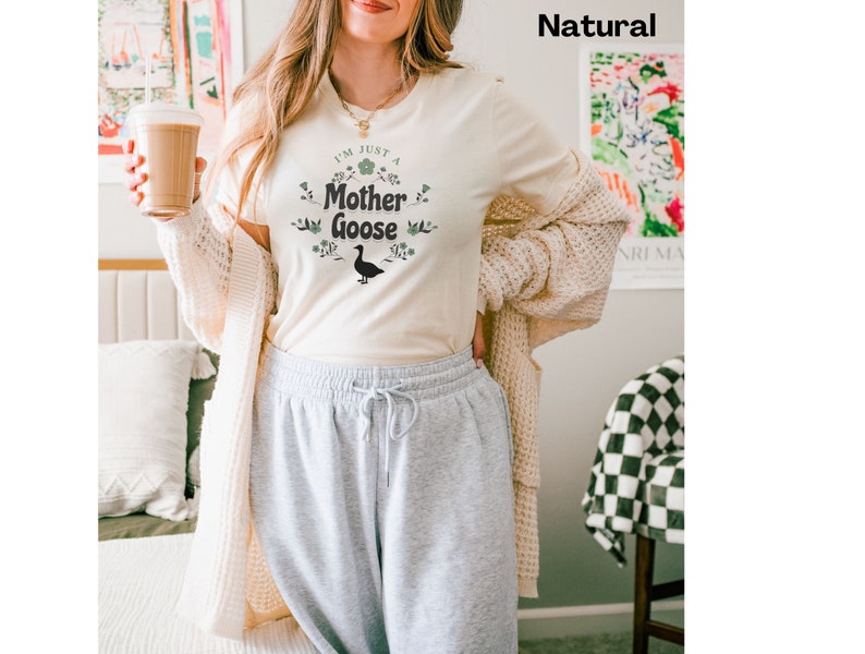 May include: Cream-colored t-shirt with the text "I'm Just a Mother Goose" in a floral design, featuring a black goose silhouette. The shirt is paired with gray sweatpants and a cream-colored cardigan.