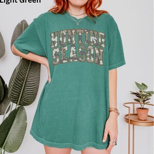 K&ouml;nnte beinhalten: Hellgr&uuml;nes T-Shirt mit einem Camouflage-Druck der Worte "Hunting Season" in gro&szlig;er, fetter Schrift.