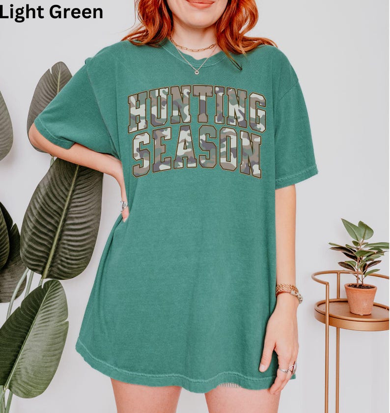 K&ouml;nnte beinhalten: Hellgr&uuml;nes T-Shirt mit einem Camouflage-Druck der Worte "Hunting Season" in gro&szlig;er, fetter Schrift.