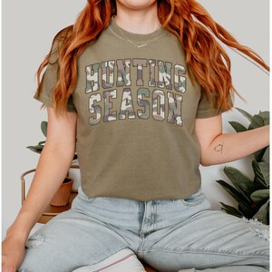 K&ouml;nnte beinhalten: Ein khakifarbenes T-Shirt mit dem Text "Hunting Season" in Tarnmuster.