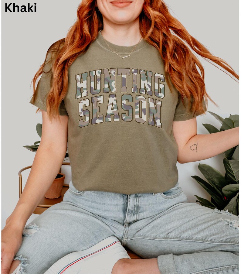 K&ouml;nnte beinhalten: Ein khakifarbenes T-Shirt mit dem Text "Hunting Season" in Tarnmuster.