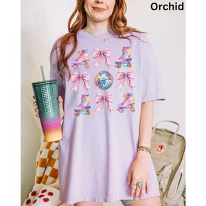 May include: A lavender t-shirt featuring a graphic print of roller skates and bows. The roller skates are rainbow coloured and the bows are pink. The background of the graphic is a disco ball.