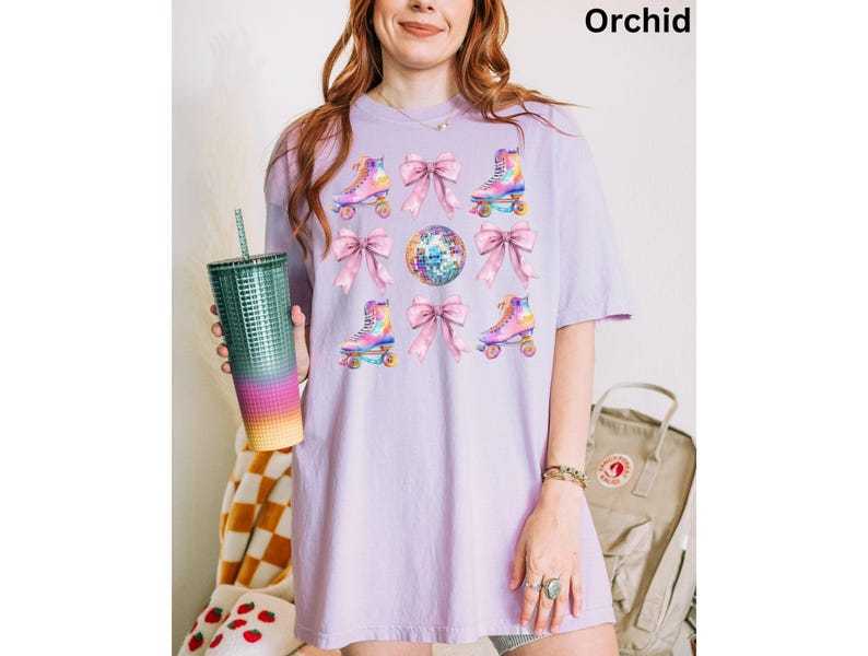 May include: A lavender t-shirt featuring a graphic print of roller skates and bows. The roller skates are rainbow coloured and the bows are pink. The background of the graphic is a disco ball.