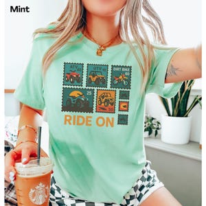 May include: Mint green t-shirt featuring a postage stamp design with various off-road vehicles, including ATVs and dirt bikes. The text "Ride On" is printed in orange below the stamps. The word "Mint" is printed in the top left corner.