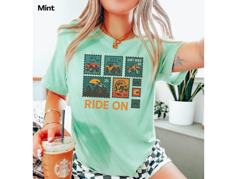 May include: Mint green t-shirt featuring a postage stamp design with various off-road vehicles, including ATVs and dirt bikes. The text "Ride On" is printed in orange below the stamps. The word "Mint" is printed in the top left corner.