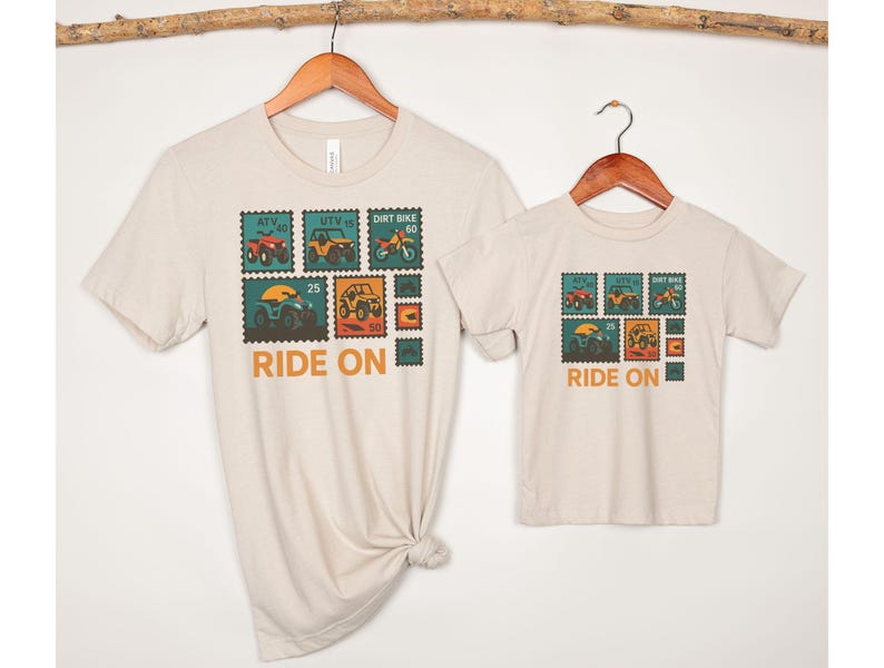 May include: Beige t-shirts, one adult and one child size, hanging on wooden hangers. Each shirt features a colorful graphic of off-road vehicles designed as postage stamps, with the words "RIDE ON" in orange.