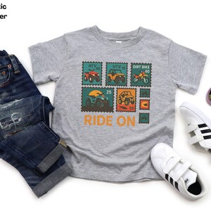 May include: Athletic heather gray t-shirt with a colorful graphic of off-road vehicles on postage stamp designs. The shirt has the words "RIDE ON" in orange. The image also includes blue jeans and white sneakers.