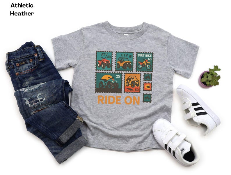 May include: Athletic heather gray t-shirt with a colorful graphic of off-road vehicles on postage stamp designs. The shirt has the words "RIDE ON" in orange. The image also includes blue jeans and white sneakers.