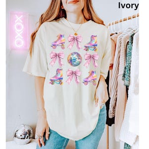 May include: A white t-shirt featuring a colourful roller skate and disco ball design. The design incorporates pink bows and iridescent roller skates. The text "Ivory" is visible in the top right corner of the image.