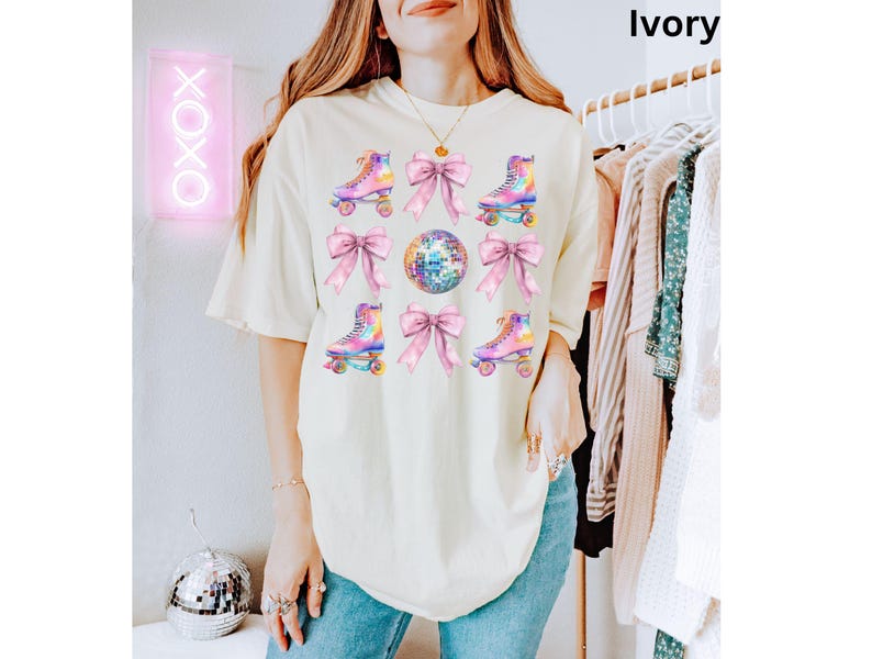 May include: A white t-shirt featuring a colourful roller skate and disco ball design. The design incorporates pink bows and iridescent roller skates. The text "Ivory" is visible in the top right corner of the image.