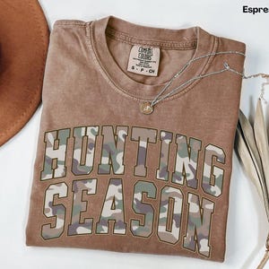 K&ouml;nnte beinhalten: Ein braunes T-Shirt mit einem Tarnmuster, auf dem "Hunting Season" steht.