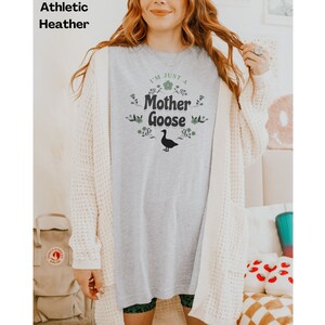 May include: Athletic heather gray t-shirt with the text "I'M JUST A Mother Goose" in green and black. The design includes floral accents and a goose silhouette. The model is wearing a cream-colored cardigan.
