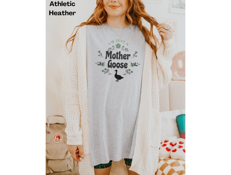 May include: Athletic heather gray t-shirt with the text "I'M JUST A Mother Goose" in green and black. The design includes floral accents and a goose silhouette. The model is wearing a cream-colored cardigan.