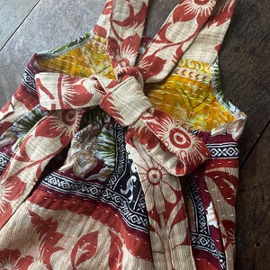 May include: A sleeveless romper featuring a multi-coloured floral and geometric pattern in shades of rust, yellow, and black. The garment includes shoulder straps and a tie-front closure at the chest, finished with visible white running stitch detailing.