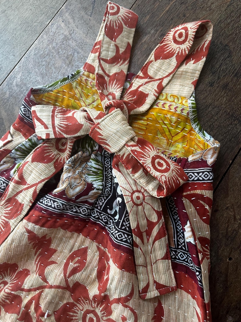 May include: A sleeveless romper featuring a multi-coloured floral and geometric pattern in shades of rust, yellow, and black. The garment includes shoulder straps and a tie-front closure at the chest, finished with visible white running stitch detailing.