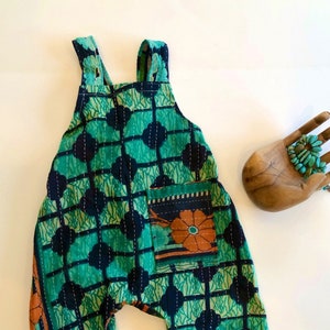 May include: A sleeveless romper featuring a vibrant green and dark blue grid pattern with white stitched detailing. The garment includes a single front pocket adorned with a floral design in orange and teal, and wide shoulder straps for a secure fit.