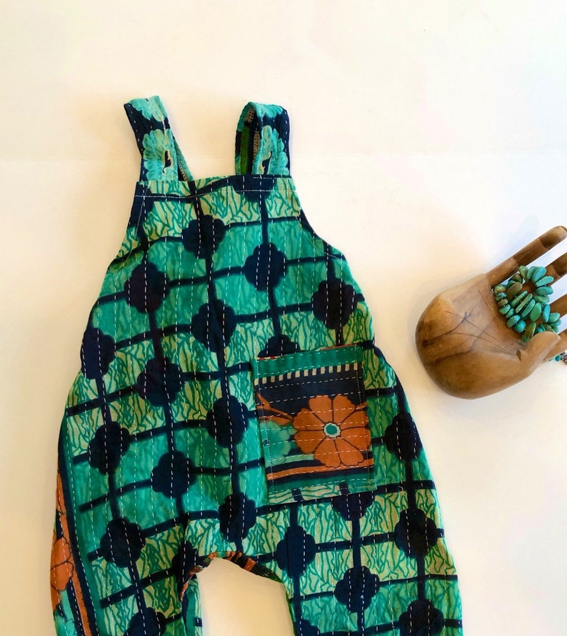May include: A sleeveless romper featuring a vibrant green and dark blue grid pattern with white stitched detailing. The garment includes a single front pocket adorned with a floral design in orange and teal, and wide shoulder straps for a secure fit.