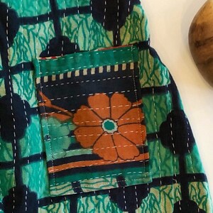 May include: A quilted fabric garment featuring a green and black grid pattern with white running stitch details. A rectangular patch pocket is attached to the front, displaying a large orange flower graphic against a dark background.