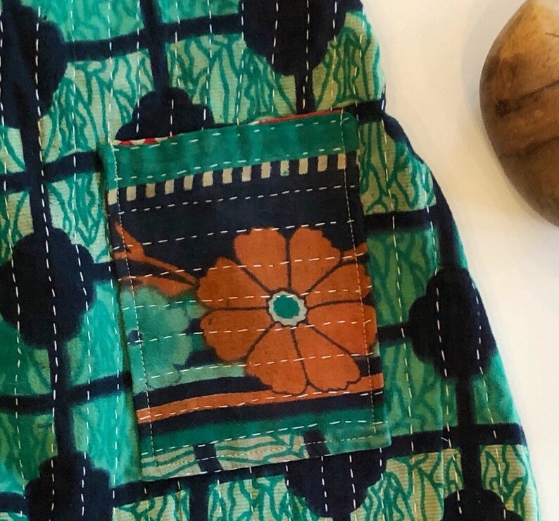 May include: A quilted fabric garment featuring a green and black grid pattern with white running stitch details. A rectangular patch pocket is attached to the front, displaying a large orange flower graphic against a dark background.