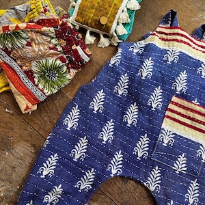 May include: A blue jumpsuit with a white leaf pattern, a striped chest pocket, and elastic cuffs. Beside it are folded trousers with a multi-coloured floral print and a rectangular mustard yellow quilted pouch with tassels and metallic circular accents.