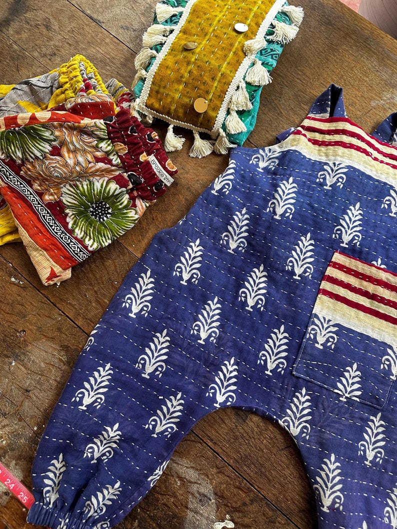 May include: A blue jumpsuit with a white leaf pattern, a striped chest pocket, and elastic cuffs. Beside it are folded trousers with a multi-coloured floral print and a rectangular mustard yellow quilted pouch with tassels and metallic circular accents.