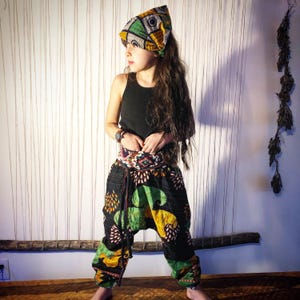May include: A black vest top paired with loose-fitting, drop-crotch trousers featuring a vibrant, multi-coloured geometric and floral pattern in shades of green, yellow, and black, complemented by a matching patterned headscarf tied around the head.