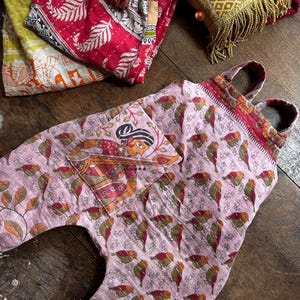 May include: A light pink quilted overall featuring a repeating pattern of small birds and a large front pocket with a printed illustration of a person playing a stringed instrument. The garment has two shoulder straps and visible white stitching.