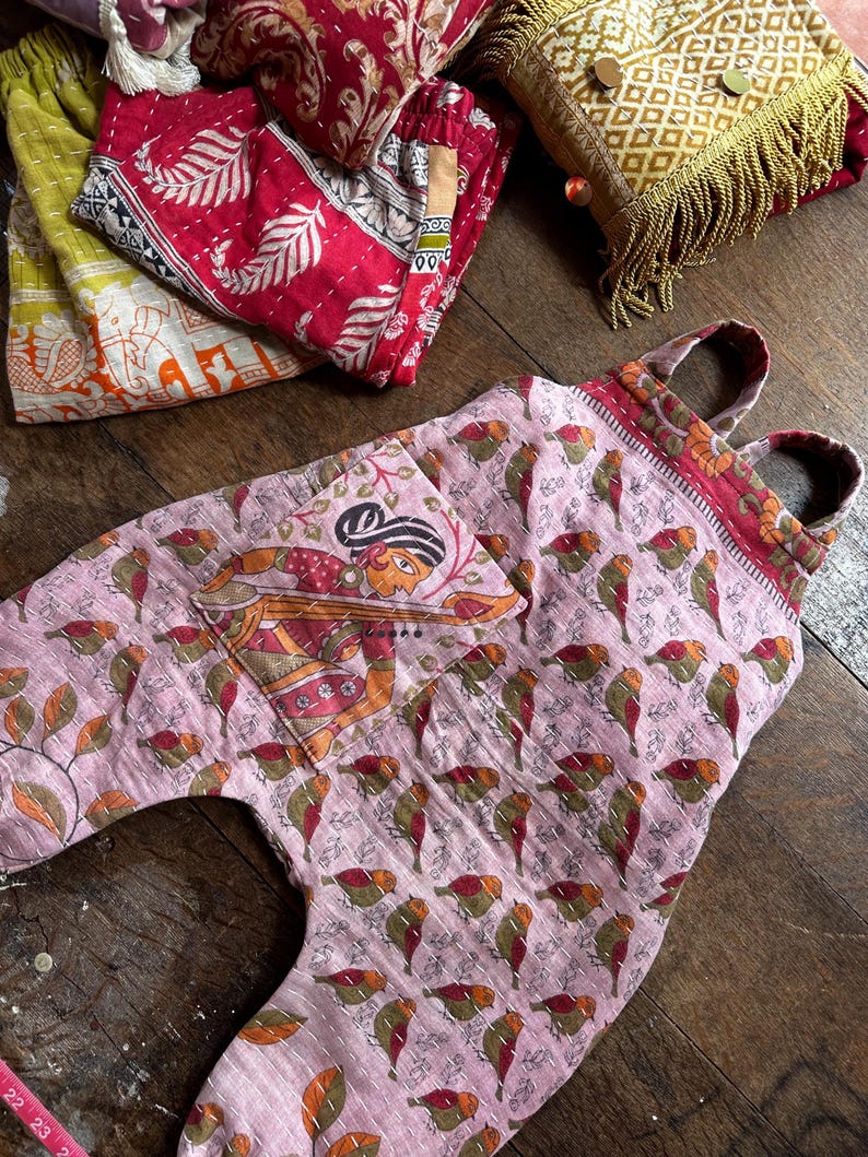 May include: A light pink quilted overall featuring a repeating pattern of small birds and a large front pocket with a printed illustration of a person playing a stringed instrument. The garment has two shoulder straps and visible white stitching.