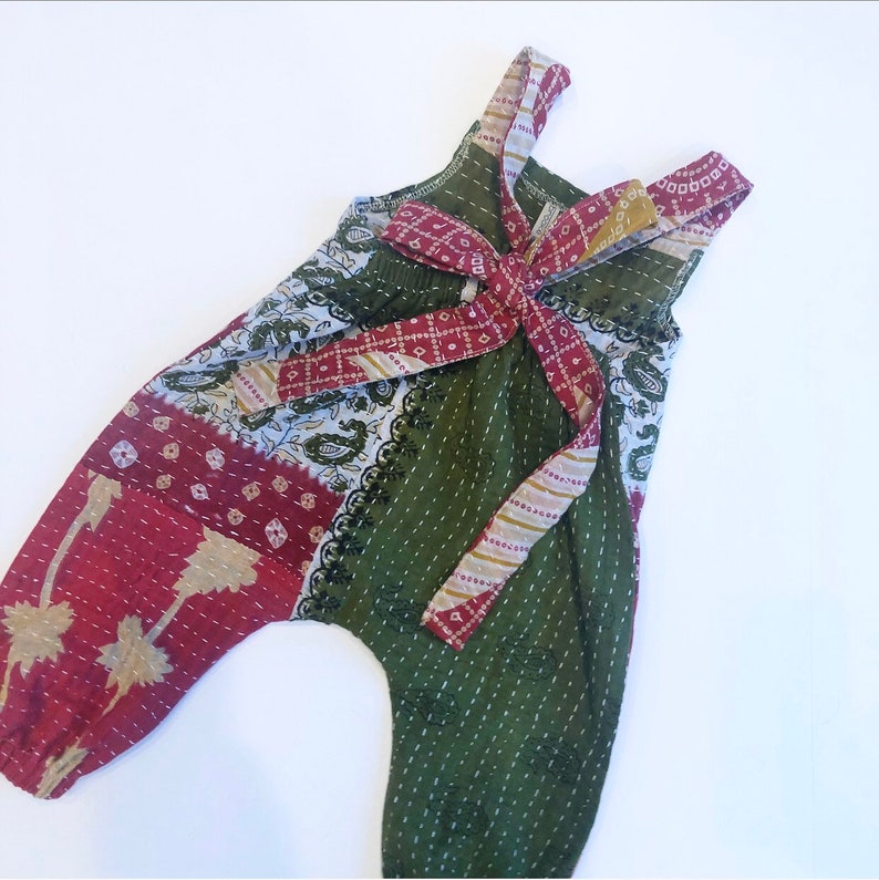 May include: A sleeveless romper featuring a patchwork design with olive green, deep red, and white fabric panels. The garment includes a tie-front detail at the chest, elasticated leg openings, and visible white running stitch embroidery throughout.