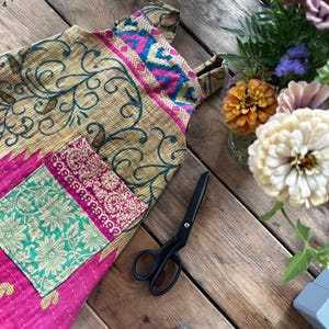 May include: A quilted apron featuring a patchwork design with a mustard yellow base, dark green swirling patterns, and a bright pink lower section. It includes a rectangular pocket with a green and pink floral print and black metal scissors.