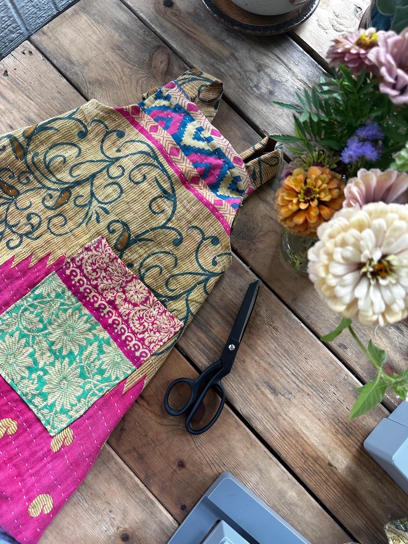 May include: A quilted apron featuring a patchwork design with a mustard yellow base, dark green swirling patterns, and a bright pink lower section. It includes a rectangular pocket with a green and pink floral print and black metal scissors.