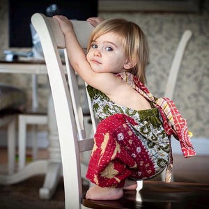 May include: A child wearing a sleeveless romper featuring a patchwork design with red, green, and white floral and geometric patterns. The garment has a tie-back closure and a gathered waist, constructed from quilted fabric with visible stitching.